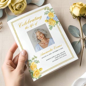 Może przedstawiać: Biały program żałobny ze złotymi obramowaniami i kwiatowymi detalami, trzymany w dłoni. Program zawiera zdjęcie kobiety i tekst "Celebrating the life of Amanda Evans". Żółte róże i zieleń otaczają program.