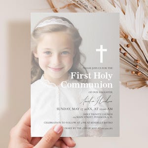 Puede incluir: Una tarjeta de invitación a la Primera Comunión sostenida por una mano. La tarjeta presenta una foto de una niña, una cruz blanca y el texto "Primera Comunión" y los detalles del evento. La tarjeta está decorada con flores secas.