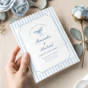 Puede incluir: Un programa de boda con un diseño de rayas azules y blancas. El programa presenta los nombres "Amanda & Michael" y detalles de la boda. El programa se sostiene en una mano, con rosas azules y vegetación en el fondo.