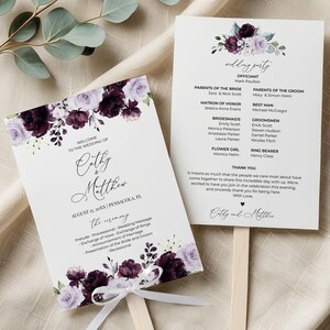 May include: Wedding fan featuring a floral design in purple and white. The fan displays the couple's names, wedding date, and ceremony information. A white ribbon is tied at the base of the fan.