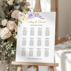 Puede incluir: Un plano de asientos de boda blanco sobre un caballete de madera, con letras doradas que dicen "Amy & David" y "PLEASE FIND YOUR SEAT". Decoraciones florales en morado, blanco y amarillo adornan la parte superior. La fecha y el lugar están en la parte inferior.