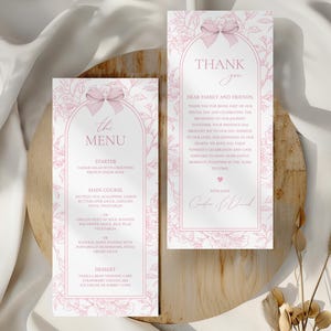 Puede incluir: Dos tarjetas de papelería de boda rosas con detalles florales y lazos. Una tarjeta dice "The Menu" con una lista de platos. La otra tarjeta dice "Thank You" con un mensaje. Ambas tarjetas están sobre una superficie de madera.