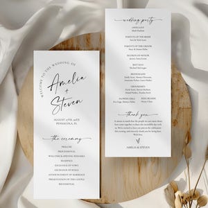 May include: Two white wedding programs with black text. The left program reads "Welcome to the Wedding of Amelia + Steven" and lists the ceremony details. The right program lists the wedding party. The programs are on a wooden surface.