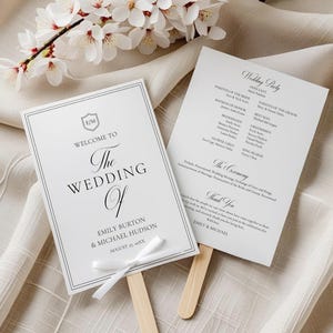May include: Two white wedding fan programs. One reads "Welcome to The Wedding Of Emily Burton & Michael Hudson". The other lists the wedding party. A white ribbon is tied to the fan's wooden stick. White flowers are in the background.