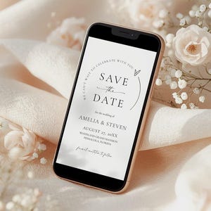 Puede incluir: Un teléfono inteligente muestra una invitación de boda con el texto "Save the Date" en una escritura elegante. La invitación incluye los nombres Amelia & Steven, la fecha 27 de agosto de 20XX y el lugar. El teléfono descansa sobre una tela suave color crema, con detalles florales.