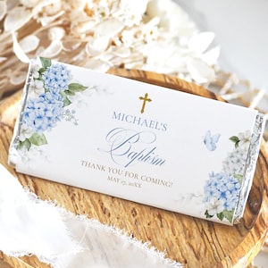 May include: A white candy bar wrapper with blue hydrangea floral design and gold cross. The text reads "Michael's Baptism" and "Thank you for coming!" with a date. A blue butterfly is also featured.