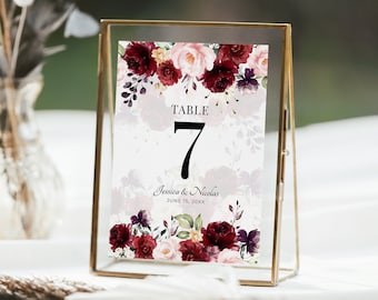 May include: A gold-framed table number card with a floral design featuring burgundy, pink, and green flowers. The card reads "Table 7" and "Jessica & Nicholas June 15, 20XX".