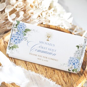 Op de afbeelding: Een witte chocoladereepverpakking met blauwe hortensia bloemaccenten en gouden tekst. De verpakking leest "Michael's First Holy Communion" met een kelk illustratie. Op een houten dienblad met een wit lint.