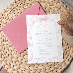 May include: A pink invitation card with floral accents and the text "Baptism & First Birthday" is placed on a woven mat, next to a pink envelope. The card includes details such as the date, time, and location.