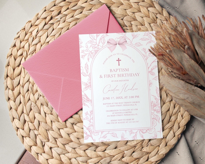 May include: A pink invitation card with floral accents and the text "Baptism & First Birthday" is placed on a woven mat, next to a pink envelope. The card includes details such as the date, time, and location.
