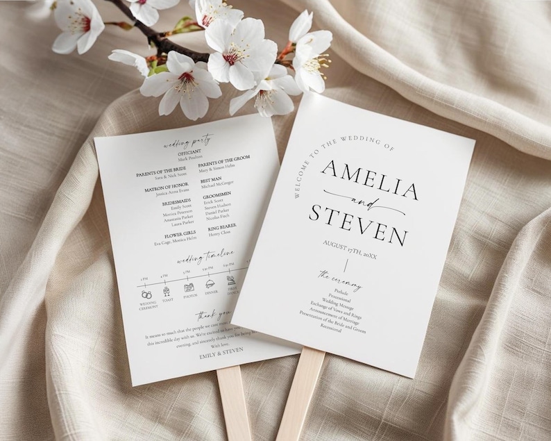 May include: Two white wedding fans with black text, one listing the wedding party and timeline, the other welcoming guests to Amelia and Steven's wedding. Cherry blossoms and a beige fabric background.