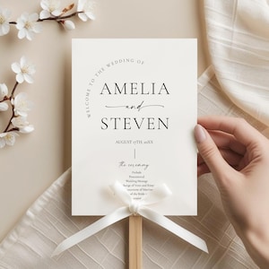May include: A wedding ceremony sign with the names "AMELIA and STEVEN" in elegant black script. The sign is held by a wooden dowel and adorned with a white ribbon. Delicate white flowers and a beige fabric are in the background.