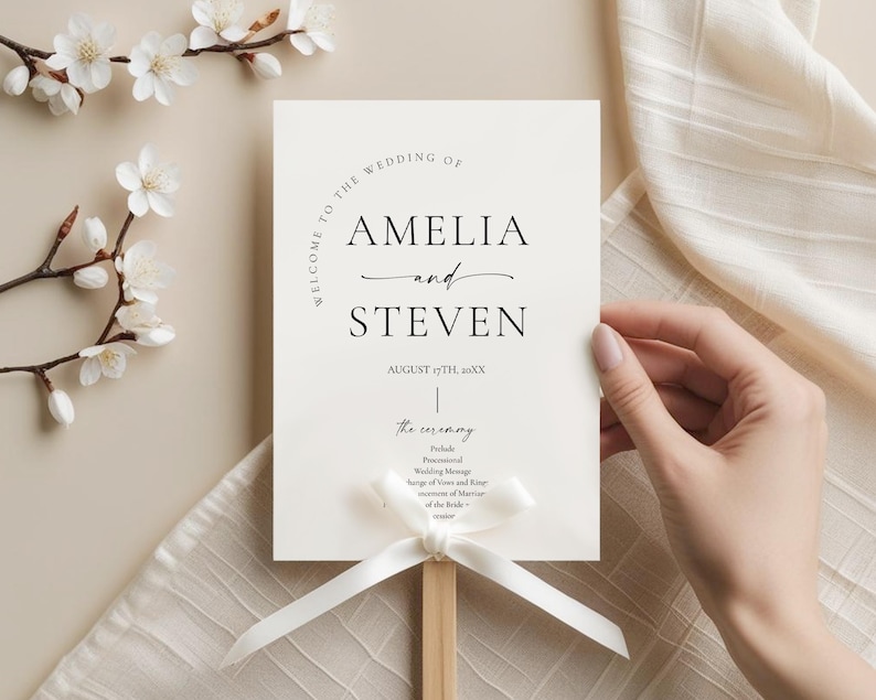 May include: A wedding ceremony sign with the names "AMELIA and STEVEN" in elegant black script. The sign is held by a wooden dowel and adorned with a white ribbon. Delicate white flowers and a beige fabric are in the background.