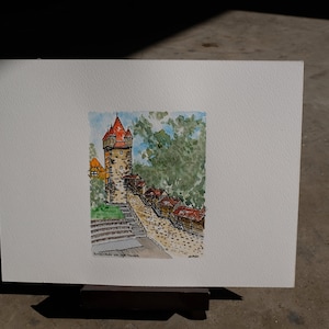 May include: Watercolour painting of a stone tower with a red roof, surrounded by trees and small buildings. The tower is on a hill with steps leading up to it. The painting is titled "Brunnenstraße und die Kirche".