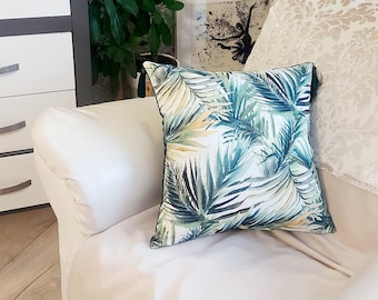 Op de afbeelding: Een witte bank met een groen en geel kussen met een tropisch bladmotief. De bank staat in een kamer met een bloemstuk, een plant en een ingelijst schilderij.
