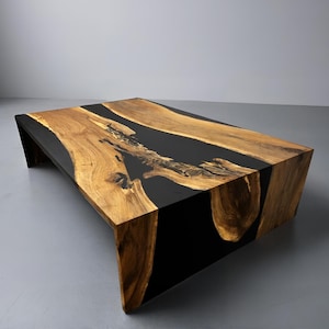 May include: A rectangular coffee table with a natural wood grain top and black resin accents. The table has a modern design with angled legs and a smooth, polished finish. The wood is a warm brown colour.
