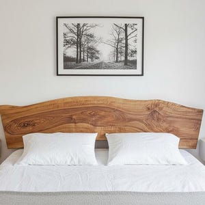 Op de afbeelding: Een slaapkamer met een houten hoofdbord met een natuurlijke, onregelmatige vorm. Het bed heeft witte lakens en kussens. Een zwart-wit landschapskunstwerk hangt boven het bed. Een kleine nachtkastje met een plant staat links.