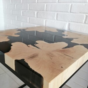 K&ouml;nnte beinhalten: Ein quadratischer Holztisch mit einer schwarzen Epoxidharzplatte. Der Tisch hat ein schwarzes Metallgestell.