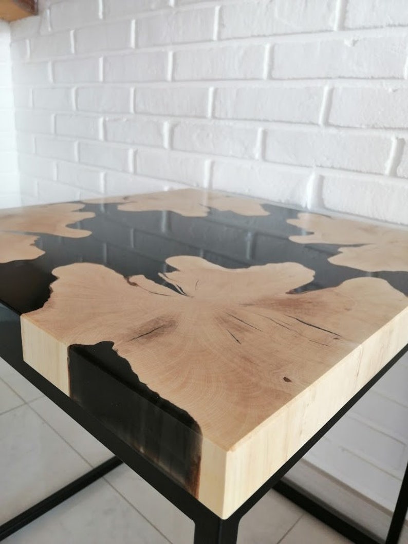 K&ouml;nnte beinhalten: Ein quadratischer Holztisch mit einer schwarzen Epoxidharzplatte. Der Tisch hat ein schwarzes Metallgestell.