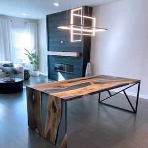 May include: A large wooden dining table with a live edge top and a gray epoxy resin inlay. The table has a black metal base with a geometric design. The table is in a modern living room with a fireplace and a chandelier.
