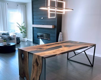 May include: A large wooden dining table with a live edge top and a gray epoxy resin inlay. The table has a black metal base with a geometric design. The table is in a modern living room with a fireplace and a chandelier.