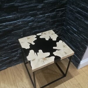 K&ouml;nnte beinhalten: Ein quadratischer Beistelltisch aus Holz mit einer schwarzen Epoxidharzeinlage. Der Tisch hat einen schwarzen Metallrahmen.