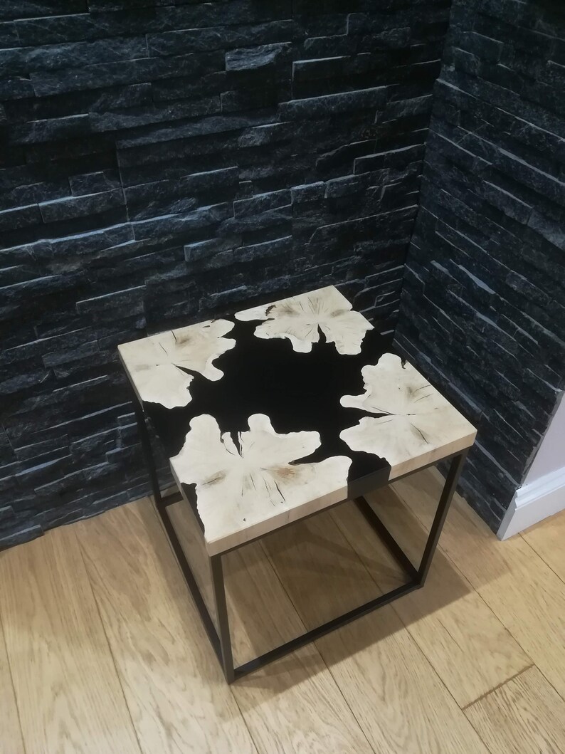 K&ouml;nnte beinhalten: Ein quadratischer Beistelltisch aus Holz mit einer schwarzen Epoxidharzeinlage. Der Tisch hat einen schwarzen Metallrahmen.