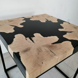 K&ouml;nnte beinhalten: Ein quadratischer Beistelltisch mit einer Tischplatte aus schwarzem Epoxidharz und Holz. Der Tisch hat ein schwarzes Metallgestell.
