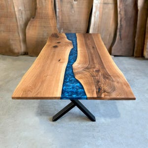 May include: A large wooden dining table with a blue resin river running down the center. The table has a black metal X-shaped base.