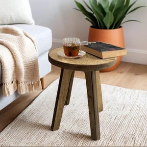 May include: A small wooden stool with a round top and three legs. The stool is painted a light brown color and has a natural wood finish. A cup of tea and a book are sitting on the stool.