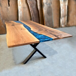 May include: A wooden dining table with a blue resin inlay. The table has a live edge and a black metal X-shaped base.