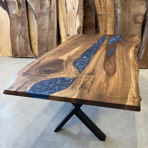May include: A large wooden dining table with a live edge top and a blue epoxy river running through the center. The table is supported by a black metal X-shaped base.