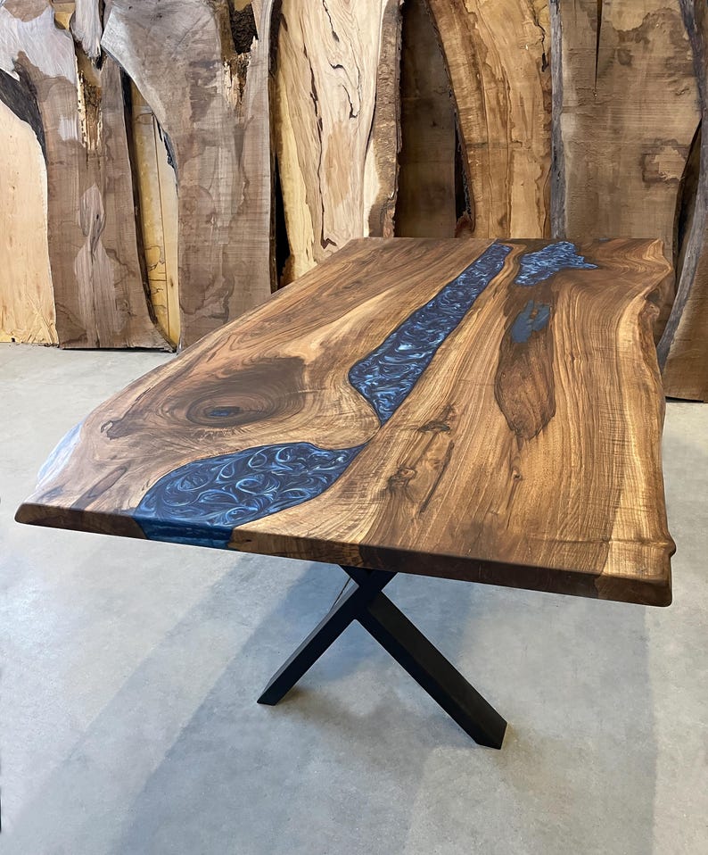 May include: A large wooden dining table with a live edge top and a blue epoxy river running through the center. The table is supported by a black metal X-shaped base.