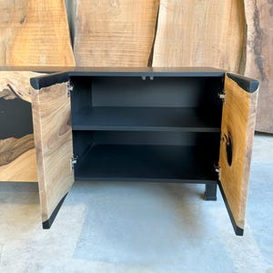 May include: A black cabinet with two wooden doors, each with a unique wood grain pattern and a black epoxy resin inlay. The cabinet has two shelves inside and is open, revealing the interior.