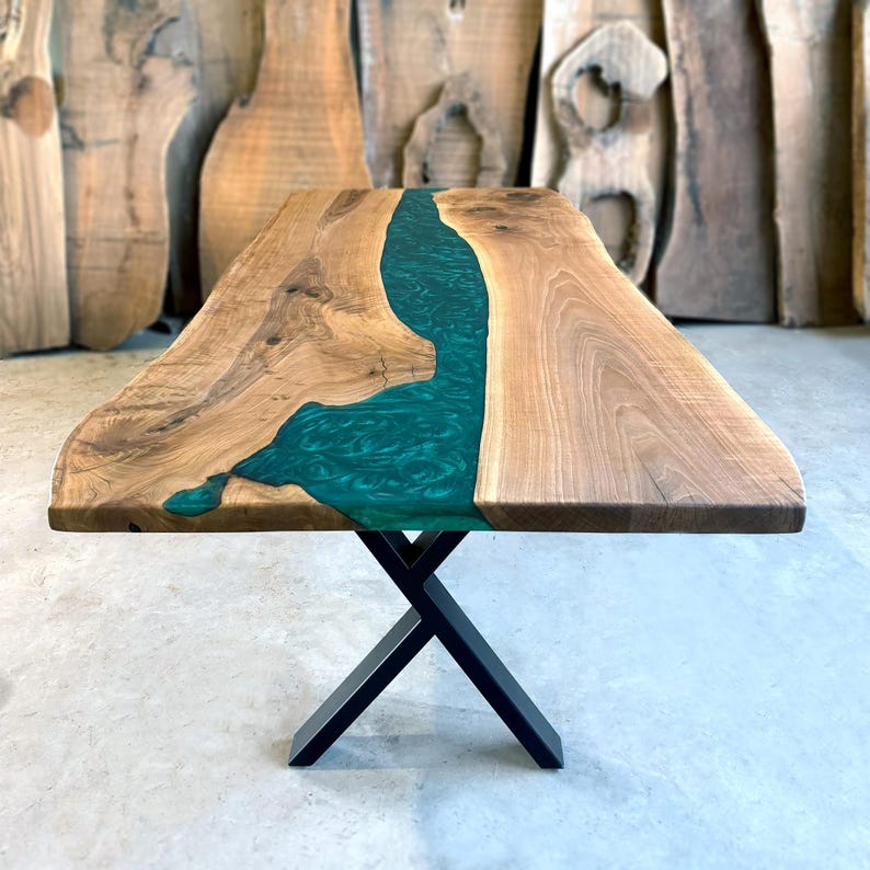 May include: A large wooden dining table with a live edge top and a green epoxy river running through the center. The table is supported by a black metal X-shaped base.