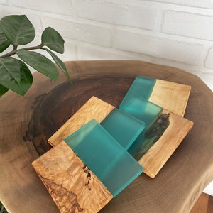 Peut inclure: Ensemble de quatre sous-verres carrés en bois avec un dessus en résine turquoise. Les sous-verres présentent un motif de grain de bois naturel et sont disposés sur une surface en bois.