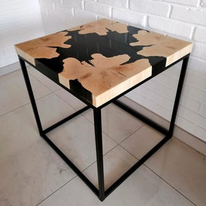 K&ouml;nnte beinhalten: Ein quadratischer Beistelltisch mit einer Tischplatte aus schwarzem Epoxidharz und Holz sowie einem schwarzen Metallgestell.