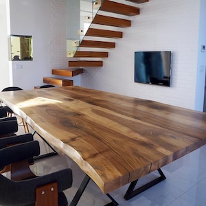 Puede incluir: Una gran mesa de comedor de madera con borde vivo, acabado en madera natural y patas de metal negro. La mesa está ubicada en un comedor moderno con una pared de ladrillo blanco y un televisor de pantalla plana montado en la pared.