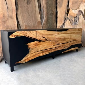 May include: A black and wood cabinet with three doors. The doors are made of wood with black epoxy resin filling in the gaps. The cabinet has black legs and is sitting on a concrete floor.