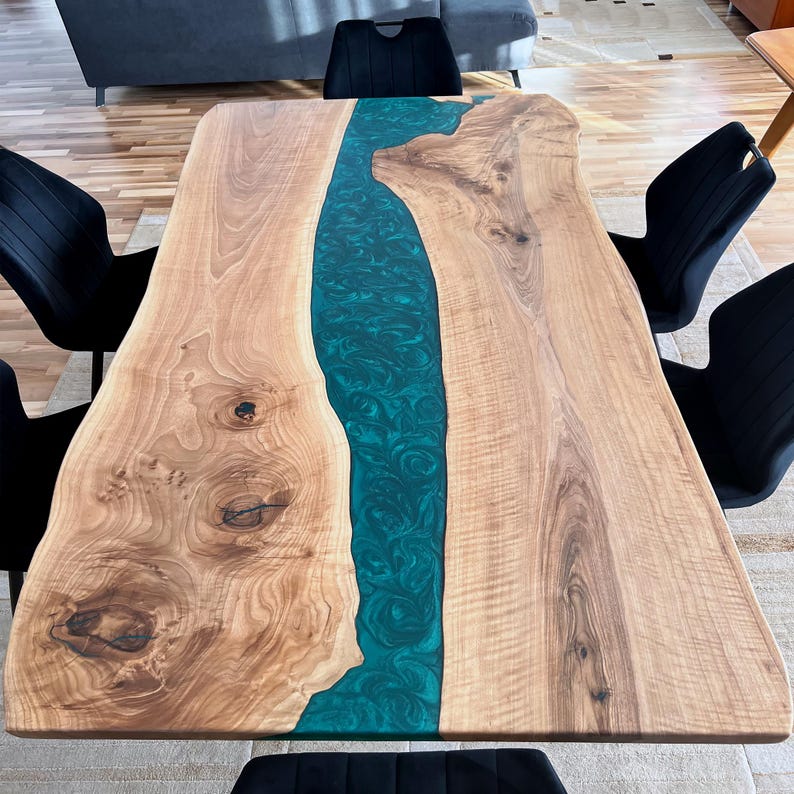 May include: A large wooden dining table with a live edge and a teal blue epoxy river running down the center. The table is surrounded by black chairs.