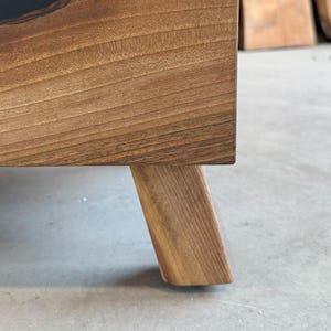 May include: Close-up of a wooden table leg with a natural wood finish. The leg is square and has a slightly rounded edge. The table leg is made of solid wood and has a natural grain pattern.