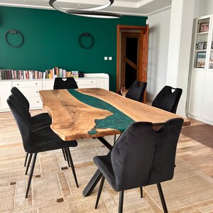 May include: A large wooden dining table with a green epoxy river running through the center. The table has a black metal base and is surrounded by black velvet chairs. The table is set on a beige rug with a geometric pattern.