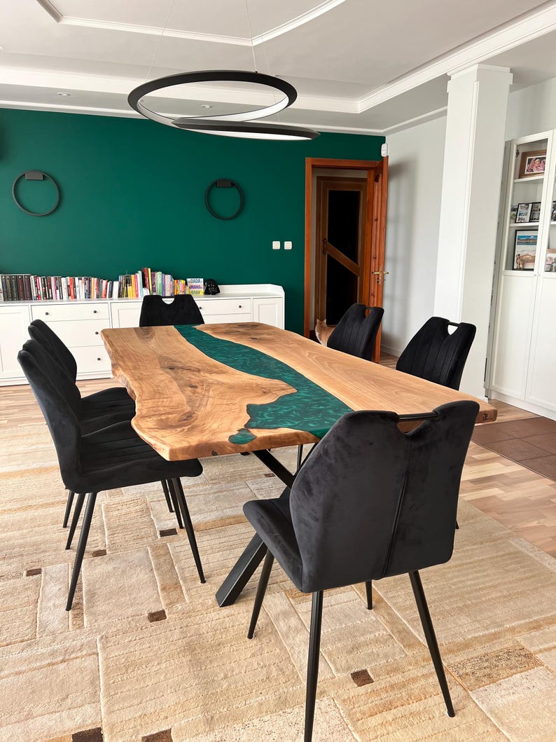 May include: A large wooden dining table with a green epoxy river running through the center. The table has a black metal base and is surrounded by black velvet chairs. The table is set on a beige rug with a geometric pattern.