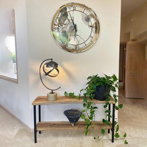 May include: A large, round wall clock with a gold-colored frame and Roman numerals hangs above a wooden console table. The table has a black metal frame and holds a decorative lamp, a potted plant, and a small black bowl. Trailing green vines add a natural touch.