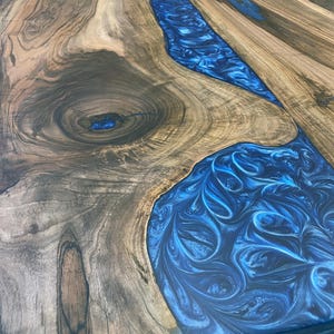 May include: A close-up of a wooden table top with a blue resin inlay. The resin has a swirling pattern and is set into the wood in a river-like shape.