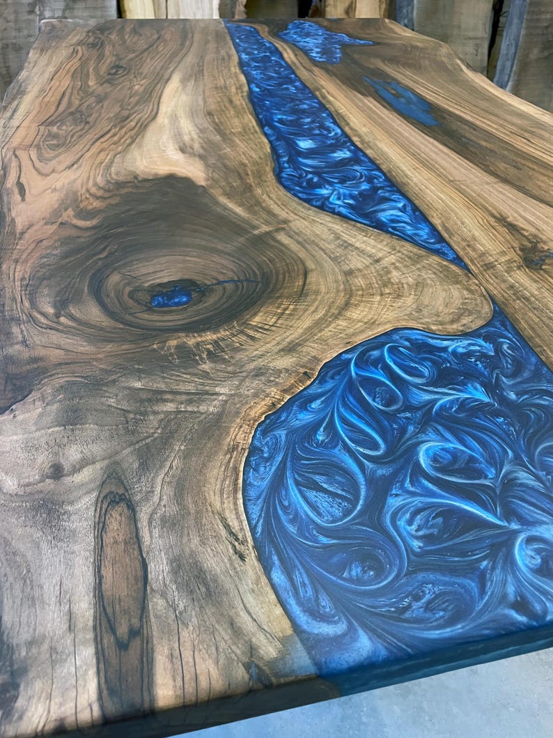 May include: A close-up of a wooden table top with a blue resin inlay. The resin has a swirling pattern and is set into the wood in a river-like shape.