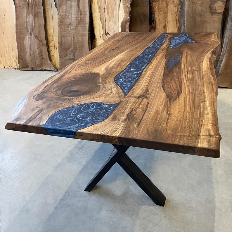 May include: A large wooden table with a live edge top and a blue epoxy river running through the center. The table is supported by a black metal X-shaped base.