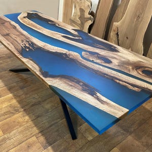 Puede incluir: Una mesa de madera con una incrustaci&oacute;n de resina epoxi azul. La mesa tiene un patr&oacute;n de veta de madera natural y la resina es lisa y brillante. La mesa est&aacute; soportada por una base de metal negro en forma de X.