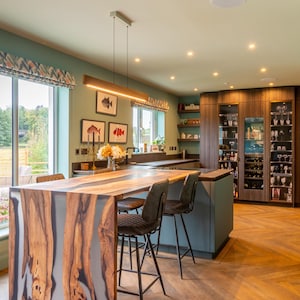 May include: A kitchen interior featuring a live-edge wood and resin breakfast bar with two dark gray bar stools. The room includes teal walls, a herringbone wood floor, a built-in wine cabinet, and three framed fish illustrations on the wall.