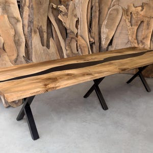 May include: A long wooden dining table with a black epoxy river running down the center. The table is supported by two black metal X-shaped legs.